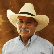 Head shot of a man in glasses with a mustache wearing a straw cowboy hat and a grey shirt.