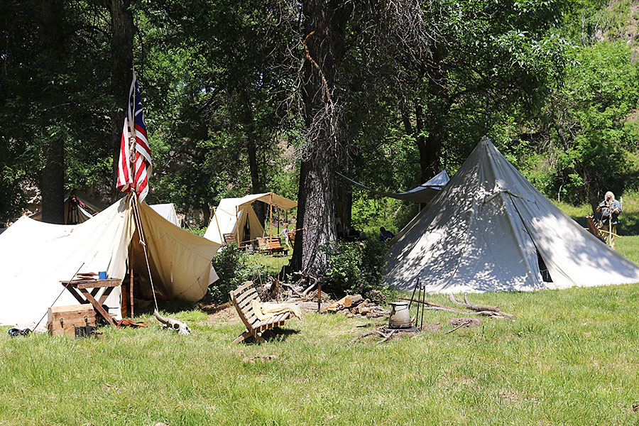 Canvas tents set up under a stand of trees. One of the tents displays an American flag.