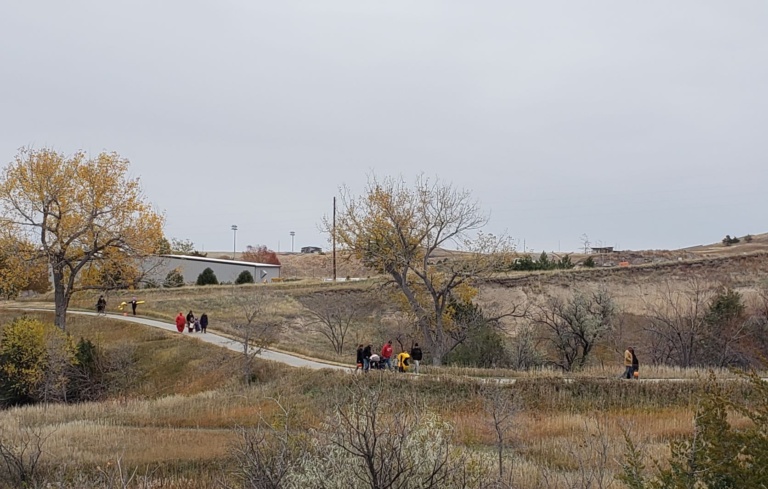 Kids in Halloween costumes hike along a paved trail flanked by trees in fall colors.