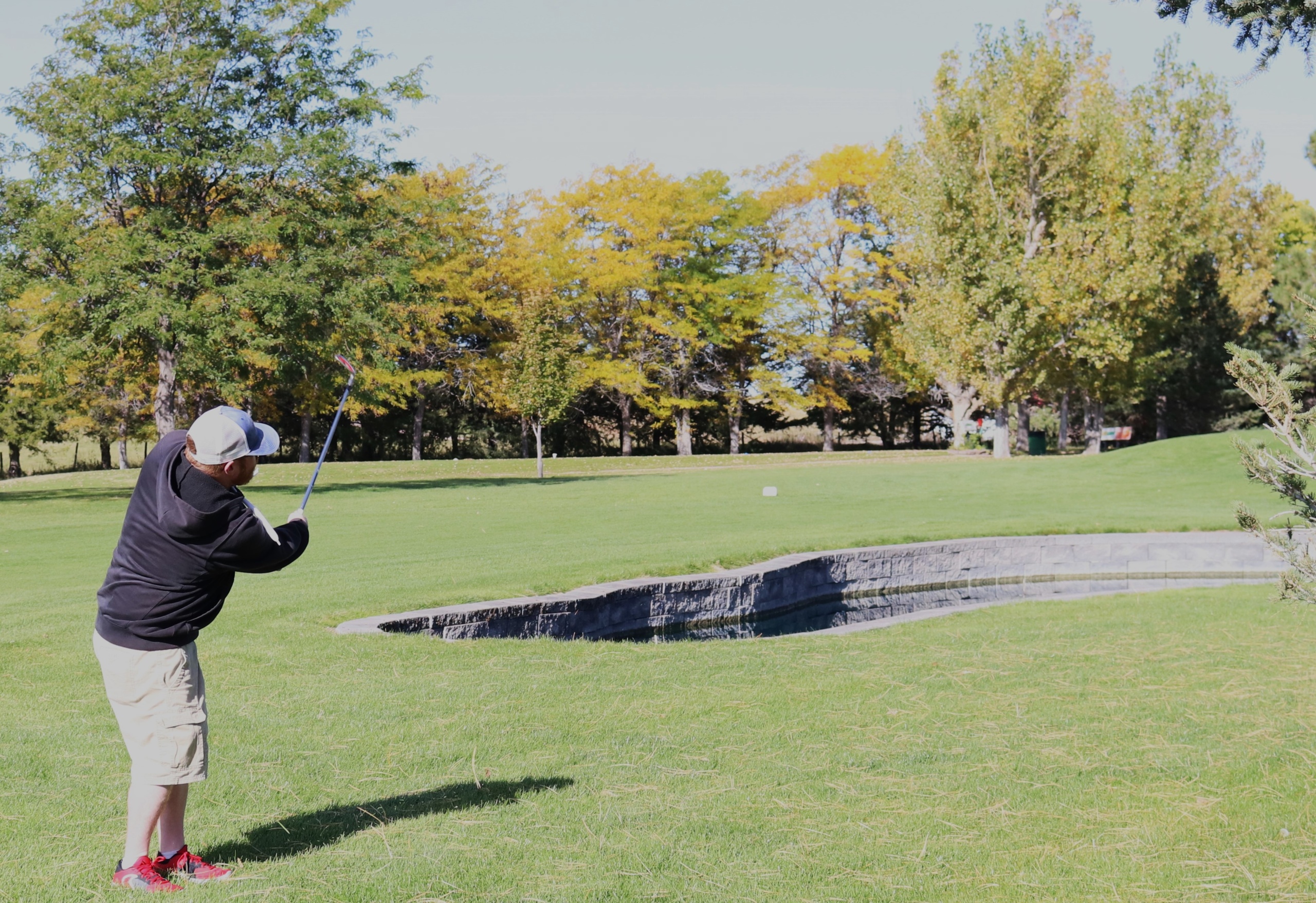 A man in brown shorts and a black sweatshirt wearing a cap swings a golf club near a water feature