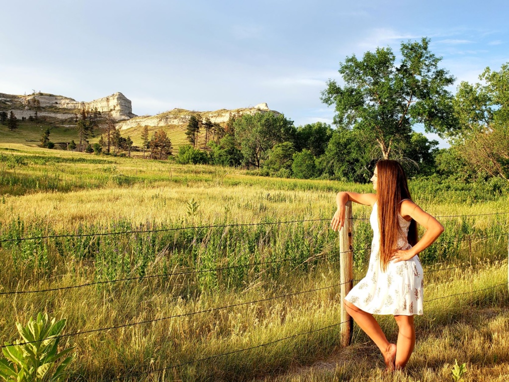 A girl with long brown hair and in a white dress stands in the green grass and gazes out on the buttes.