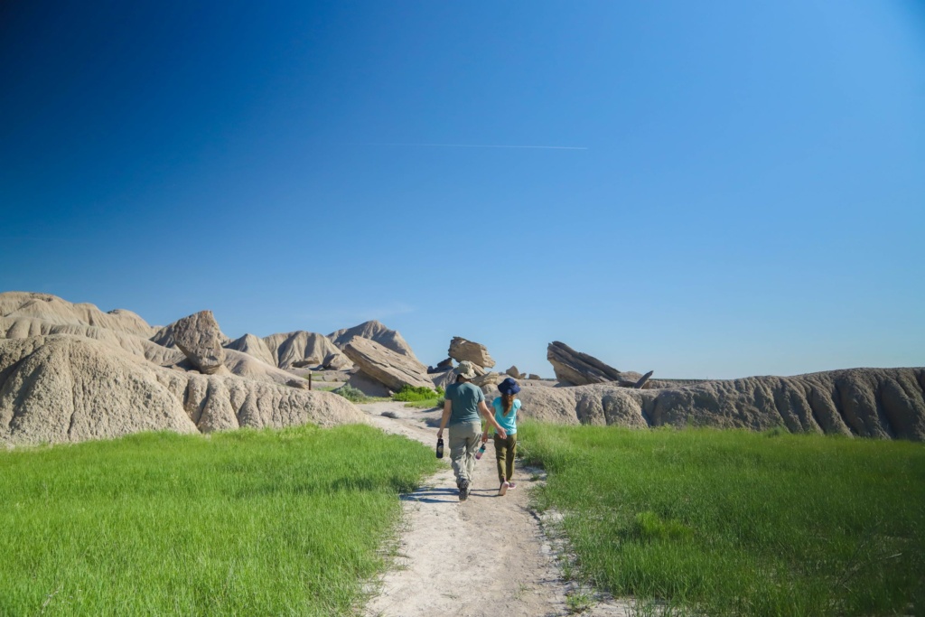 A mother and daughter wearing hats walk down a gravel trail surrounded by green grass toward large rock formations.