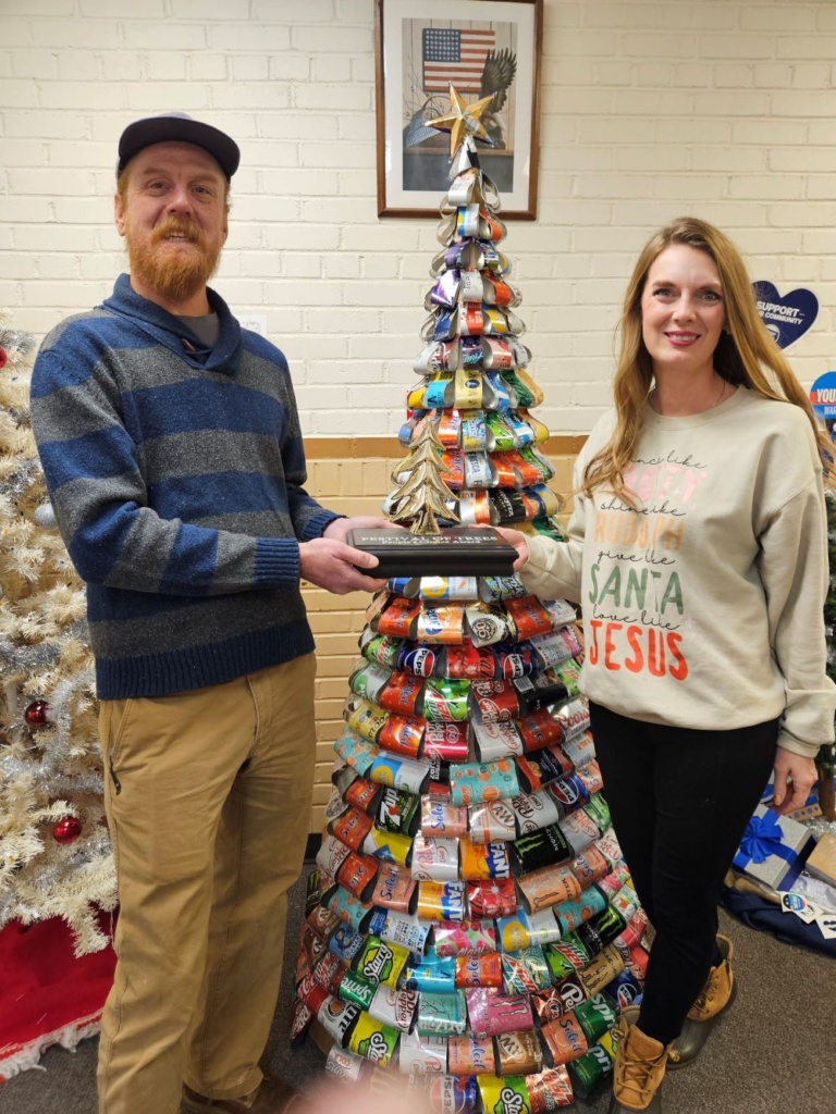 A man wearing a cap and a woman with long hair hold a trophy shaped like a Christmas tree in front of a tree made of aluminum cans.