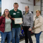 Three women and a man stand next to each other, and the man is holding an award certificate.