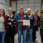 Six women stand in a row, and the third woman from the left holds an award certificate.