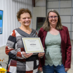 Two women stand next to each other, and the woman on the left holds an award certificate.