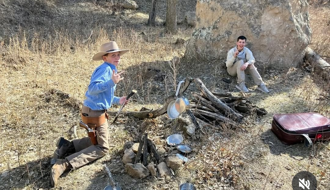 Two young men sit on the ground around the beginnings of a campfire.