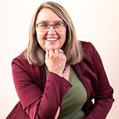 A woman with shoulder length brown hair in a burgandy jacket and green shirt and glasses rests her chin on her right hand in a headshot.