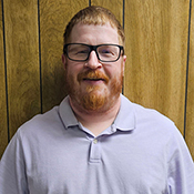 A man in a purple shirt and glasses stands against a brown wall for a head and shoulders shot.