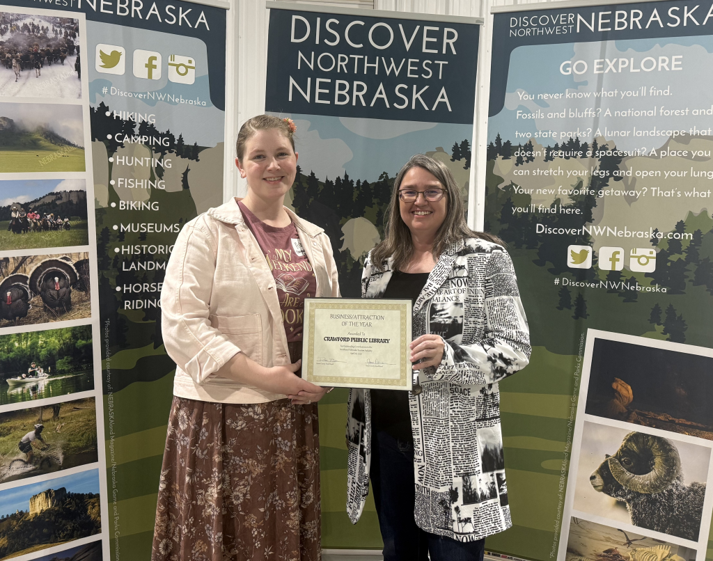 A woman in a dress and a woman in a blazer pose in front of banners that say Discover Northwest Nebraska and hold an award certificate.