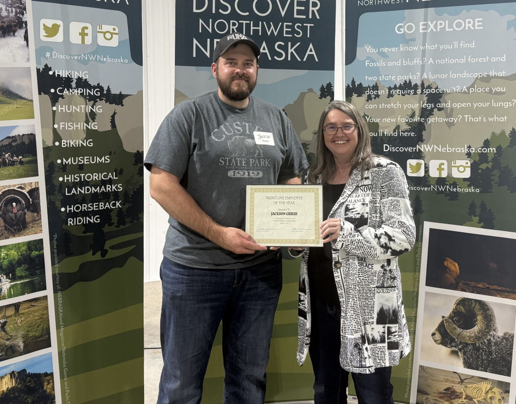 A man in a ball cap and a woman in a blazer pose in front of banners that say Discover Northwest Nebraska and hold an award certificate.