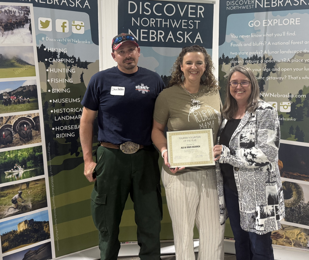 A man in a ball cap and two women pose in front of banners that say Discover Northwest Nebraska and hold an award certificate.