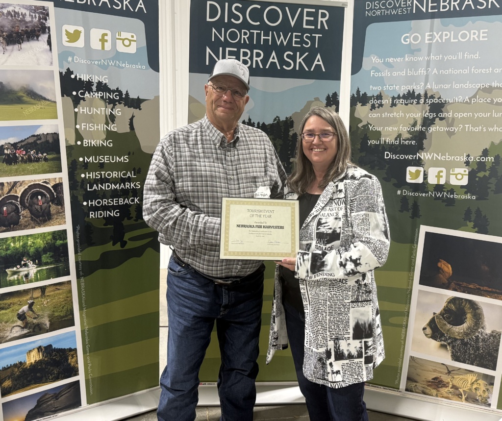 A man in a ball cap and a woman in a blazer pose in front of banners that say Discover Northwest Nebraska and hold an award certificate.