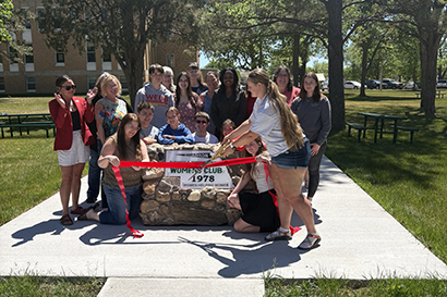 A group of women and men stand behind a stone monument with a sign that reads "Chadron Business & Professional Women's Club" and cut a red ribbon to re-dedicate it.
