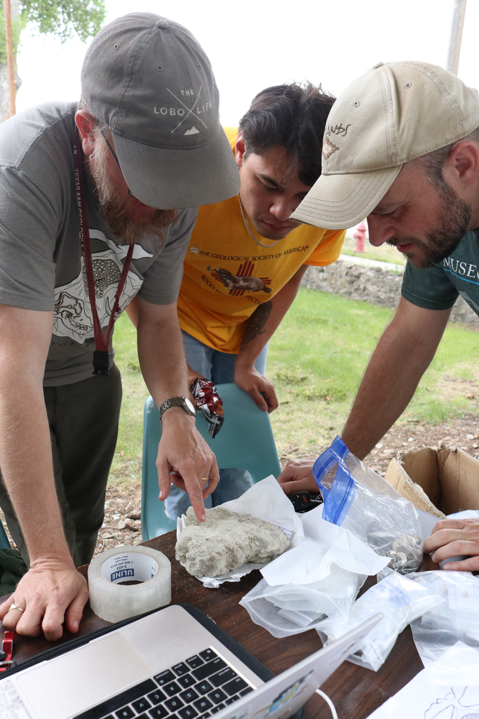 Three men, two wearing ball caps, study rocks containing fossils as they bend over a table with a laptop computer on it.