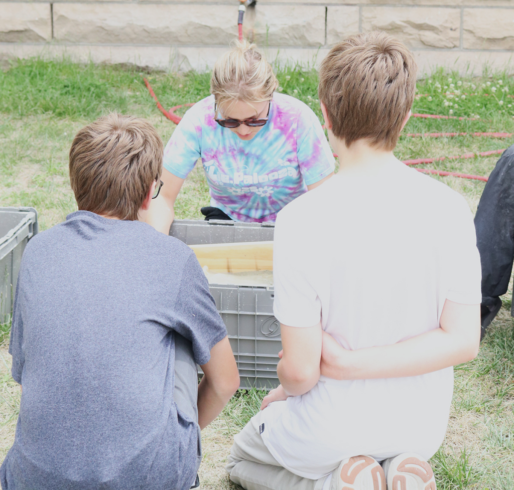 A female college student shows two boys, with their backs to the camera, how to screen for microfossils using a seive and water.