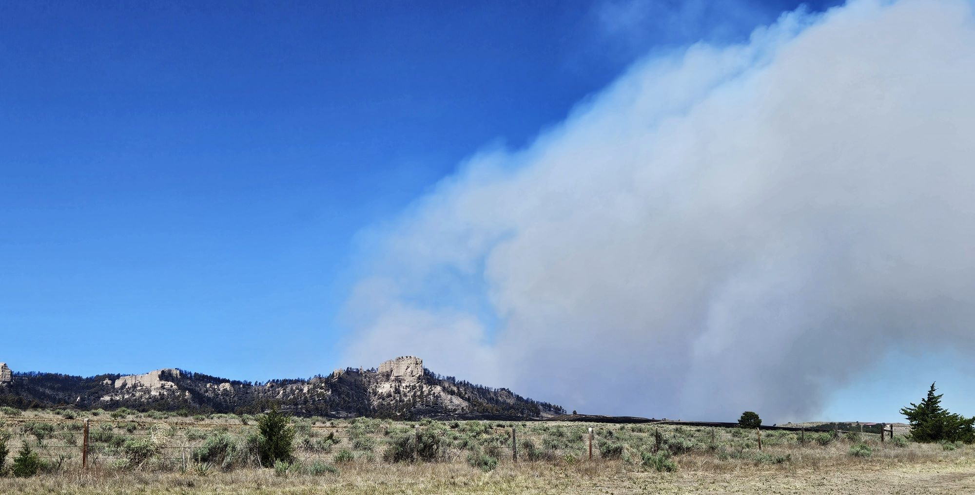 A large plume of wildfire smoke rises into the sky behind buttes.