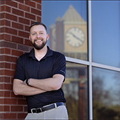 Head shot of a man with a beard wearing a black shirt leaning against a brick wall with a glass wall behind him reflecting a clock tower.